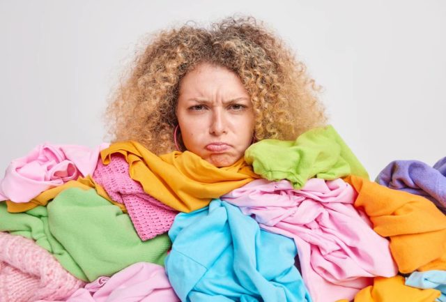 Discontent young European woman with curly hair has grumpy expression overloaded with colorful clothes upset to have much house work has to fold washed clothing isolated over white background Как правильно выбрасывать одежду
